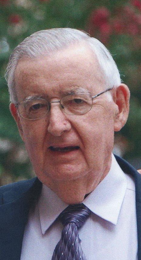 Elderly man with gray hair and glasses smiling gently, wearing a suit and tie, set against a blurred outdoor background.